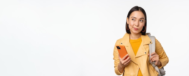 Viajante de garota asiática com mochila segurando telefone celular usando aplicativo de smartphone parecendo pensativo em pé sobre fundo branco