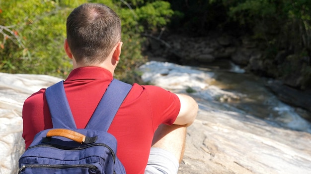 Viajante de homem de camisa polo vermelha com uma mochila azul senta-se perto de um rio de montanha