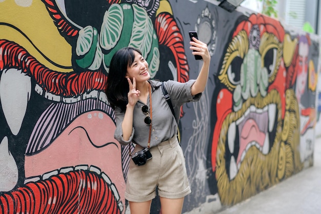 Viajante de mochila jovem asiática usando telefone celular desfrutando da parede de arte local cultural de rua Viajante verificando ruas laterais