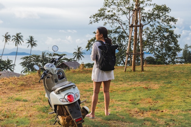 Viajante de mulher em pé perto de motocicleta na praia tropical.