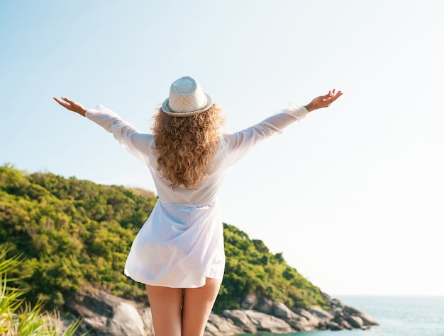 Viajante encaracolado mulher posando com levantando os braços na praia em um dia ensolarado