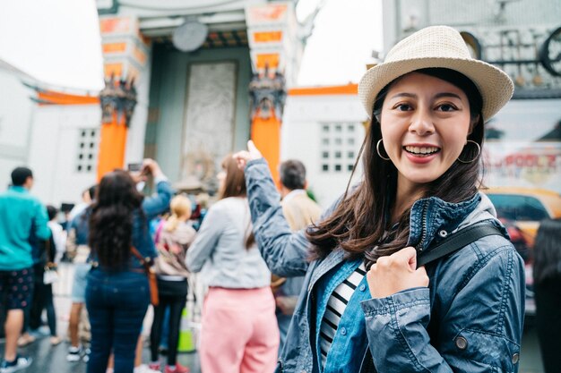 Foto viajante feminina usando chapéu de palha e animado apontando para o teatro chinês. jovem viajante cara a câmera sorrindo. mochileiro asiático mostrando a famosa paisagem urbana.