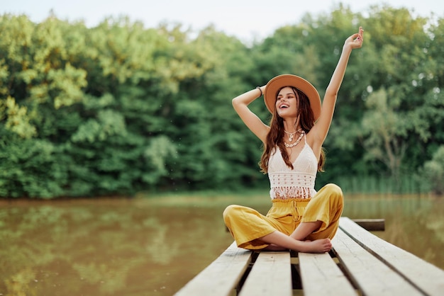 Viajante hippie ecoativista senta-se em uma ponte perto de um lago com os braços estendidos com um chapéu e sorrindo sinceramente