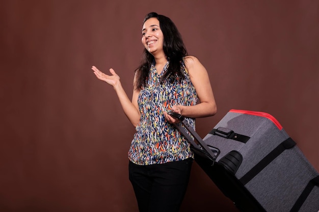 Viajante indiano sorridente em pé com mala rolante, conversando, saindo de férias. Mulher segurando bagagem sobre rodas, turista alegre conversando, carregando bagagem no aeroporto, conceito de viagem