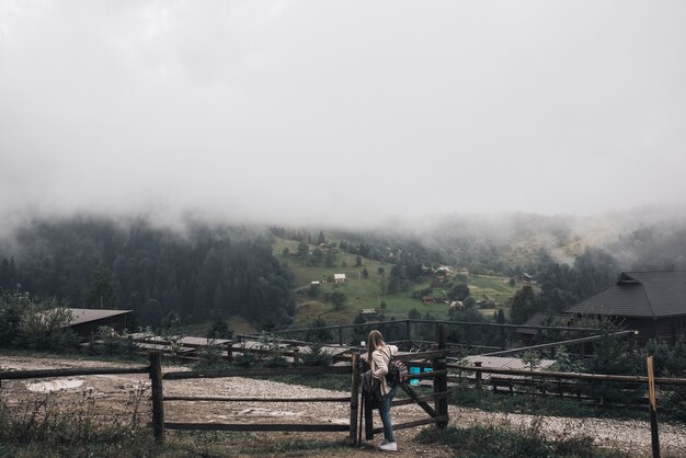 Viajante jovem e magra perto de uma cerca velha contra o fundo das altas montanhas dos cárpatos no nevoeiro