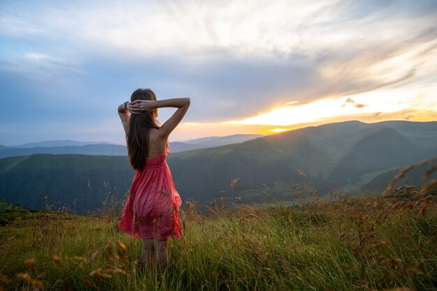 Viajante jovem mulher feliz em um vestido vermelho de pé na encosta gramada em uma noite de vento nas montanhas de verão, apreciando a vista da natureza ao pôr do sol.