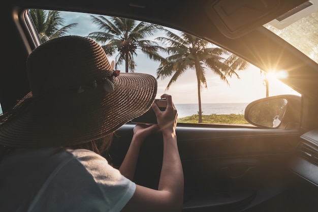 Viajante jovem olhando e tirando uma foto do pôr do sol na praia dentro do carro