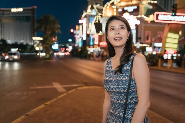 Viajante linda garota de vestido no fundo da cidade à noite com muitas luzes de outdoor brilhando em vista bokeh. turista de mulher curiosa sorrindo olhando ao redor na rua urbana no verão de las vegas nevada