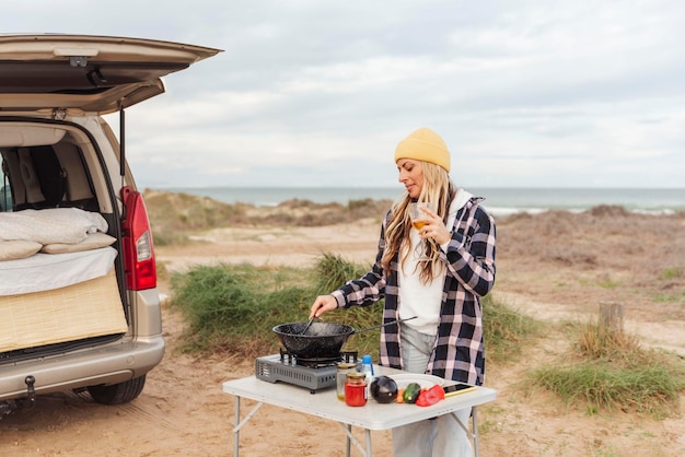 Viajante prepara comida no fogão a gás portátil em uma mesa dobrável em um local costeiro tranquilo fora de sua van enquanto usa um telefone celularConceito de viagem e estilo de vida nômade