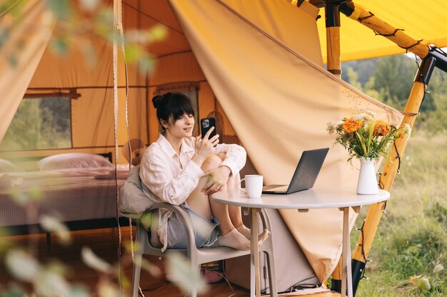 Viajante sentada ao lado da barraca de acampamento bebendo café da manhã e usando notebook trabalhando