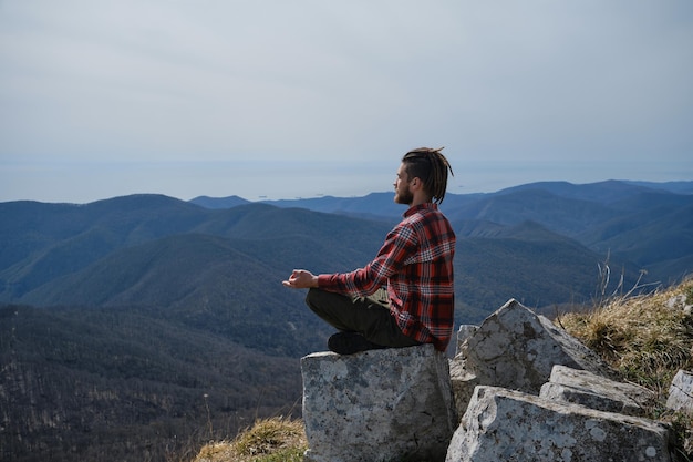 Viajante sentado em uma grande rocha nas montanhas e apreciando a vista da natureza do Cáucaso Jovem com dreadlocks medita e faz ioga fora Vista lateral Conceito de viagem e estilo de vida ativo