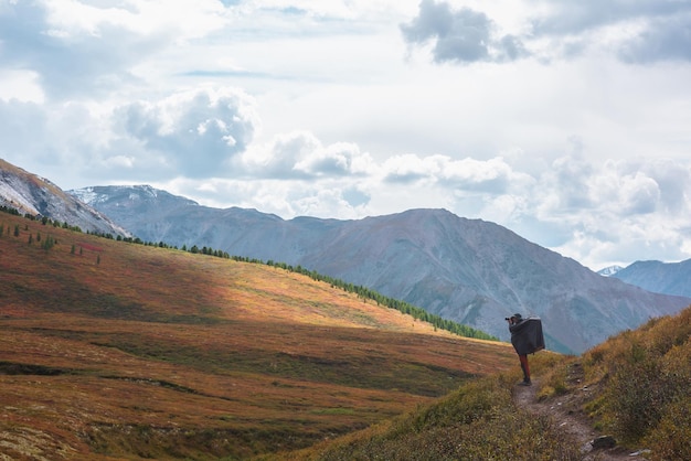 Foto viajante sozinho em impermeável com grande mochila fotografa a natureza entre a vegetação de outono desbotando em uma trilha de caminhada através de uma passagem de montanha mochileiro com câmera fotográfica filma paisagens de montanha de outono