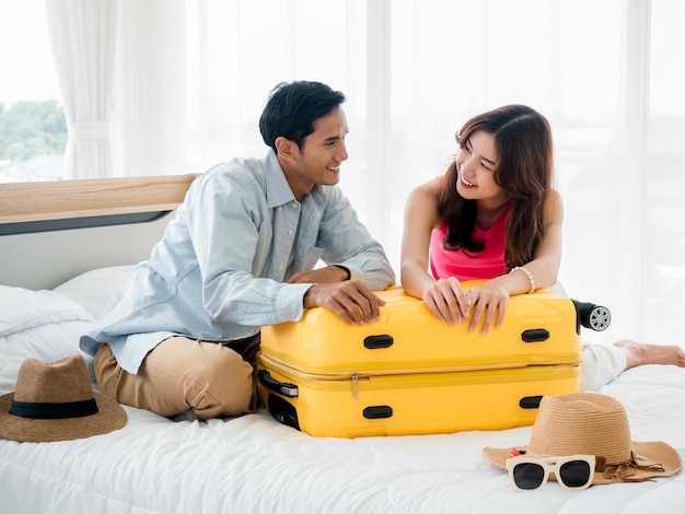 Viajantes de casal se preparando para viajar homem asiático em camisa jeans e jovem linda sentados juntos e embalando malas amarelas na cama no quarto Boas férias Férias de verão coloridas