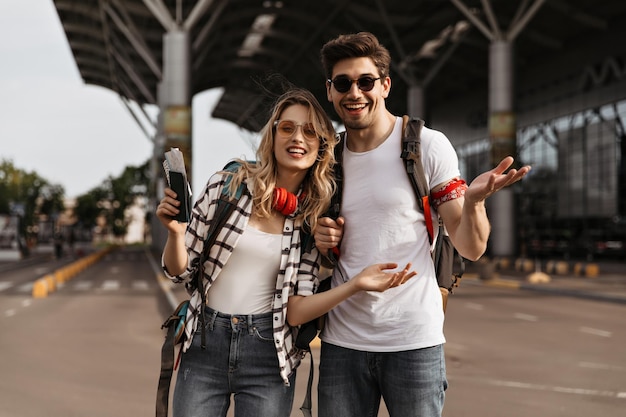 Viajantes olha para a câmera perto do aeroporto garota loira de camisa xadrez e homem de óculos escuros e camiseta branca posa com mochilas e tem passaporte