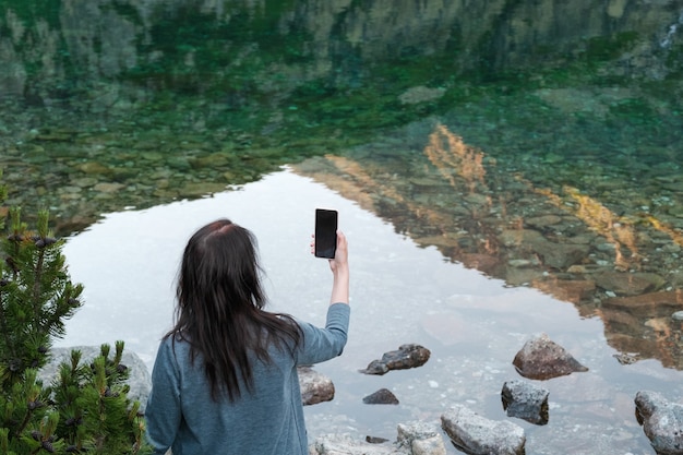 Viajar bloger joven de pie en la orilla del lago y hacer foto