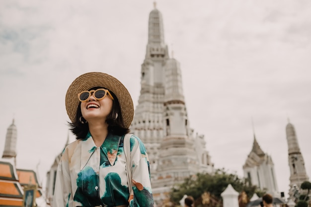 Viajar de mulher asiática com chapéu e óculos de sol está de pé na frente do templo tailandês que famoso lugar da Tailândia