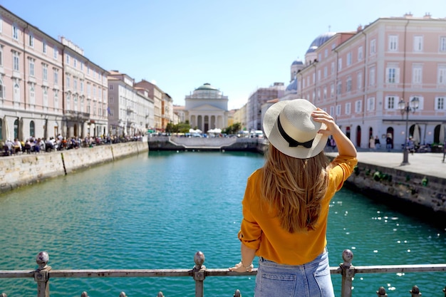 Viajar em Trieste Itália Vista traseira da menina bonita segurando o chapéu olhando para a igreja de Sant Antonio Taumaturgo no Grande Canal em Trieste Itália Bela jovem visitando a Europa