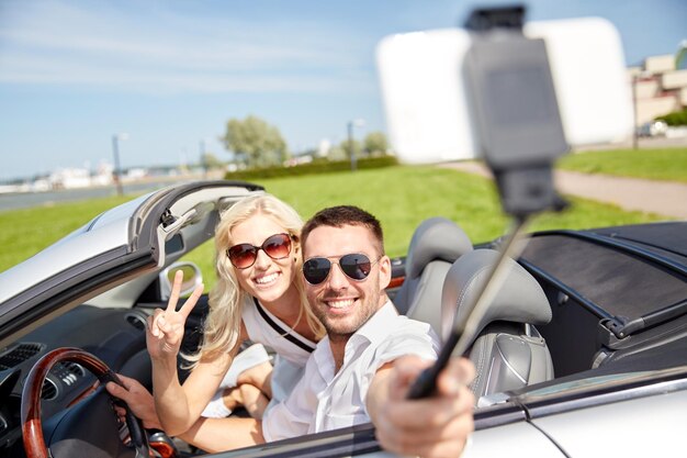 viaje por carretera, ocio, pareja, tecnología y concepto de personas - hombre y mujer felices conduciendo en coche cabriolet y tomando fotos con smartphone en selfie stick