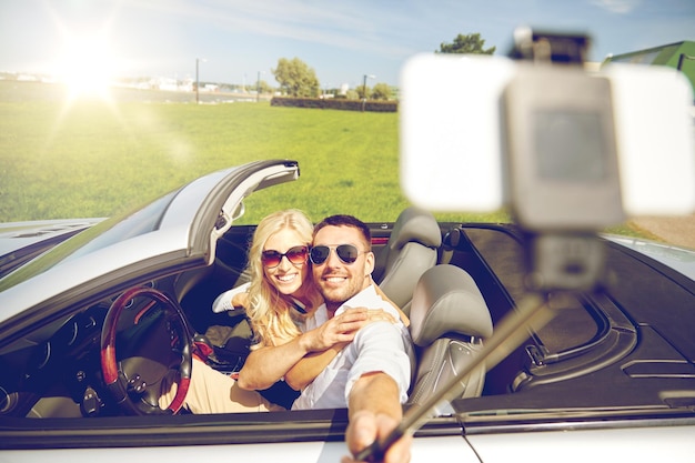 viaje por carretera, ocio, pareja, tecnología y concepto de personas - hombre y mujer felices conduciendo en coche cabriolet y tomando fotos con smartphone en selfie stick