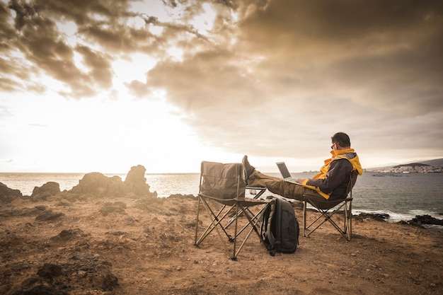 Viaje e aproveite o conceito de pessoas ao ar livre com um homem solitário trabalhando em um computador laptop conectado à Internet, sentado em frente a um incrível pôr do sol no oceano