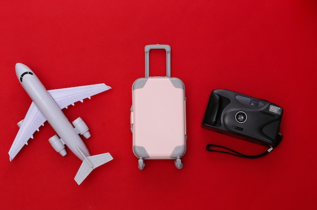 Viaje plano laical. Mini maleta de viaje de plástico, cámara, avión sobre fondo rojo. Estilo minimalista. Vista superior