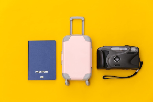 Viaje plano laical. Mini maleta de viaje de plástico, cámara y pasaporte sobre fondo amarillo. Estilo minimalista. Vista superior