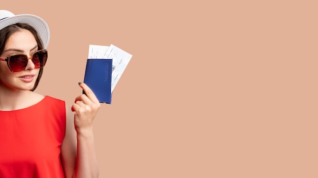 Foto viaje de verano oficina de visas mujer sonriente en vestido rojo y sombrero blanco con identificación extranjera con boletos aislados en espacio de copia beige control de pasaporte fondo publicitario