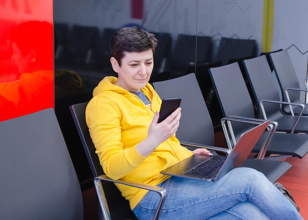 Viajera que trabaja en una laptop y usa un teléfono inteligente mientras está sentada en la terminal del aeropuerto esperando su vuelo