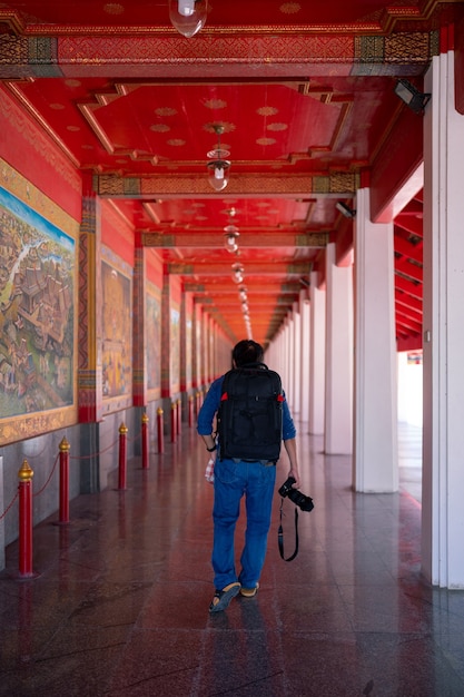 El viajero asiático de la cámara del hombre de la mochila camina en la pasarela en Wat Pa Lelai Worawihan. Pared decorada con literatura clásica Khun Chang Khun Phaen junto al templo hall, hito público en Suphanburi, Tailandia