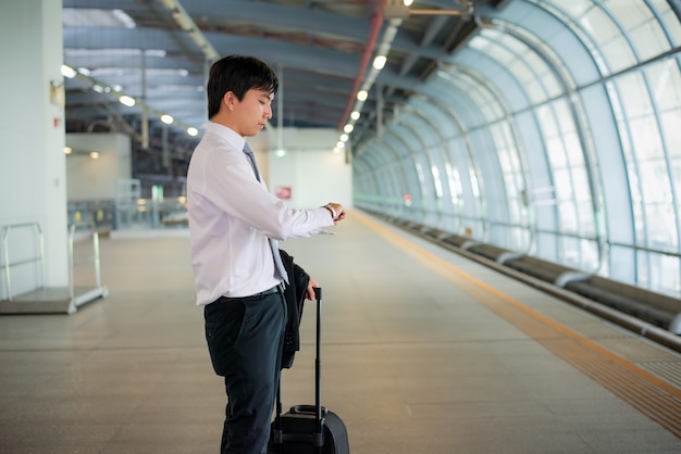 Viajero asiático joven hermoso del hombre de negocios que mira el reloj en la mano con equipaje, esperando el tren en la estación de tren, viaje y vacaciones.