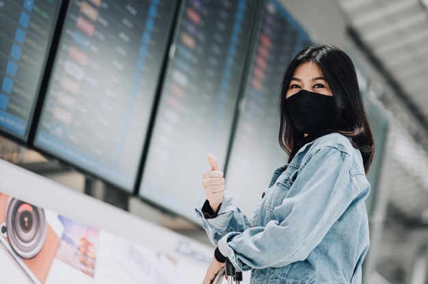 Viajero asiático con máscara protege del coronavirus mostrando el pulgar hacia arriba con confianza frente a la pantalla del tablero de información de vuelo