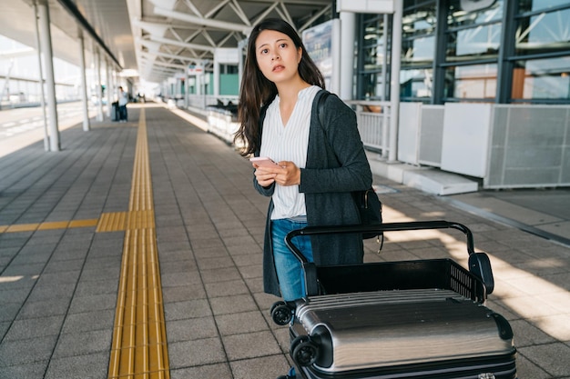 viajero asiático usando un teléfono inteligente contactando a su novio para que la recoja en el aeropuerto de kansai esperando al aire libre junto al carrito de equipaje en un día soleado. niña joven que sostiene el teléfono móvil enviando mensajes de texto
