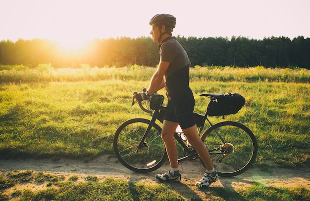 Viajero en bicicleta caminando con su bicicleta llena de bolsas y buscando el lugar para acampar al atardecer