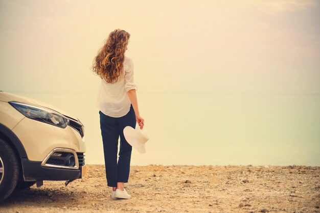 Viajero elegante feliz de la mujer joven en el camino de la playa que se sienta en el coche blanco de la cruce