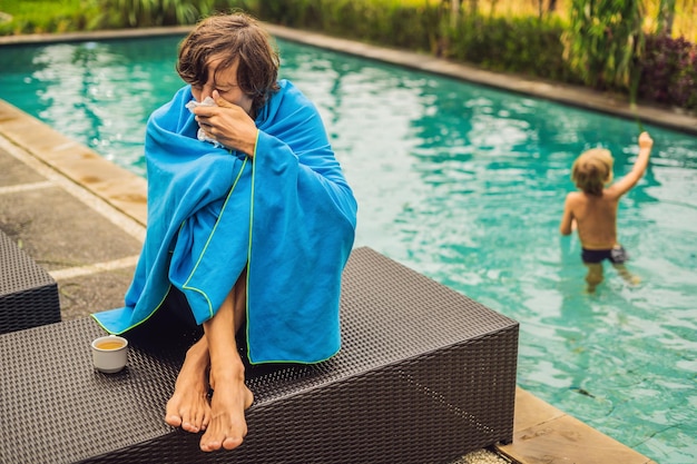 Viajero enfermo. El hombre se resfrió en las vacaciones, se sienta triste en la piscina bebiendo té y se suena la nariz con una servilleta. Su hijo está sano y nadando en la piscina. Concepto de seguro de viaje.