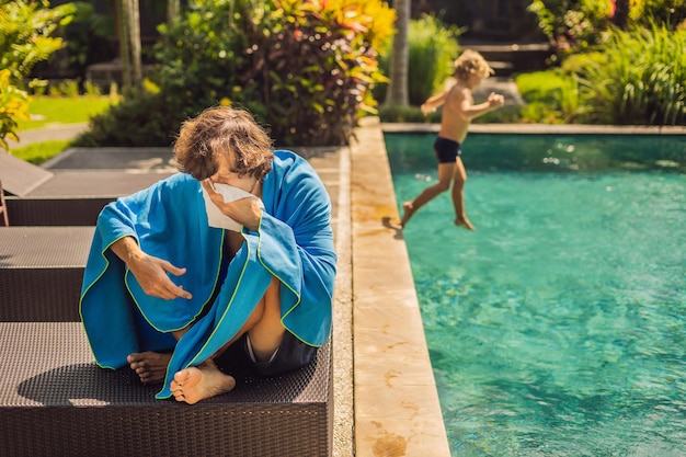 Viajero enfermo. El hombre se resfrió en las vacaciones, se sienta triste en la piscina bebiendo té y se suena la nariz con una servilleta. Su hijo está sano y nadando en la piscina. Concepto de seguro de viaje