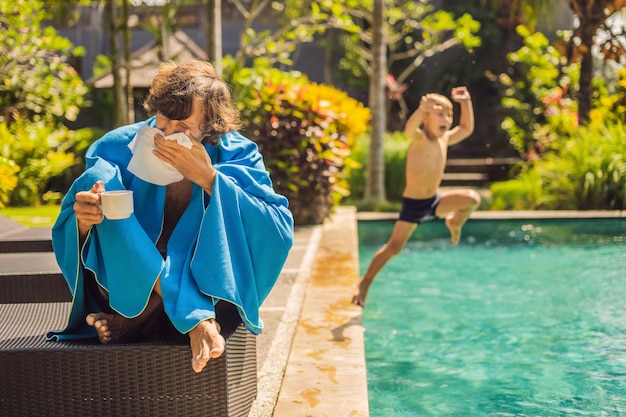 Viajero enfermo. El hombre se resfrió en las vacaciones, se sienta triste en la piscina bebiendo té y se suena la nariz con una servilleta. Su hijo está sano y nadando en la piscina. Concepto de seguro de viaje