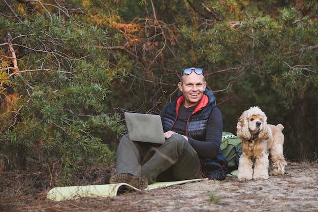 Viajero experimentado utiliza una computadora portátil con una mascota amiga durante un viaje a pie en el bosque