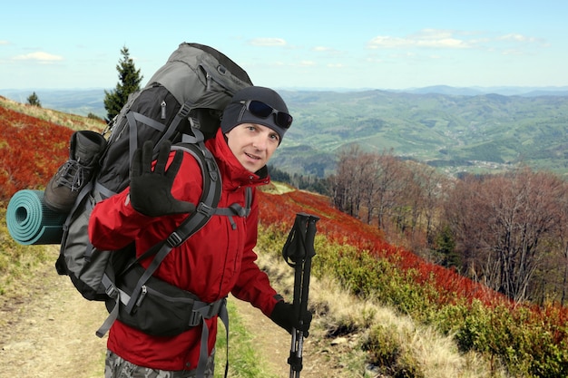 El viajero feliz equipado con una chaqueta roja en la ladera levantada en la mano de saludo