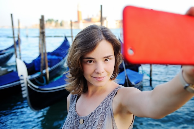 Viajero femenino joven que hace la foto del selfie usando su smartphone que se coloca cerca de la plaza de San Marco con las góndolas en el fondo en Venecia, Italia