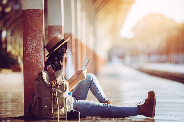 Viajero femenino con sombrero y mochila viajando con mapa de papel en la estación de tren.