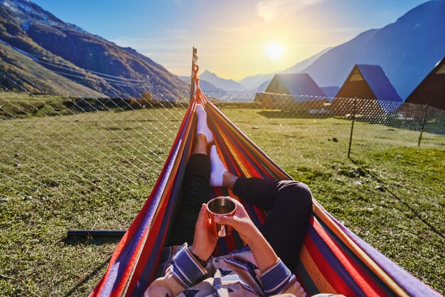 Viajero hombre tumbado en una hamaca con una taza en las manos y disfrutando de una relajante y tranquila recreación al aire libre con vistas al valle de la montaña en Georgia