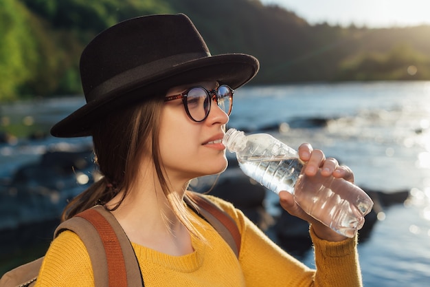 Viajero joven bebiendo agua pura de botella en el fondo del río de la naturaleza