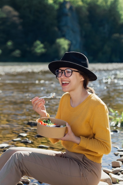 Viajero joven comer alimentos saludables cerca del río en el fondo de la naturaleza