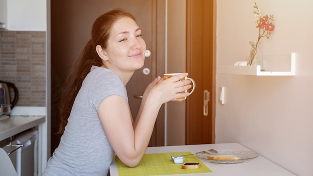 Viajero joven está desayunando en la habitación del hotel, la luz del sol. Ella está sentada a la mesa y bebiendo té, vista lateral.