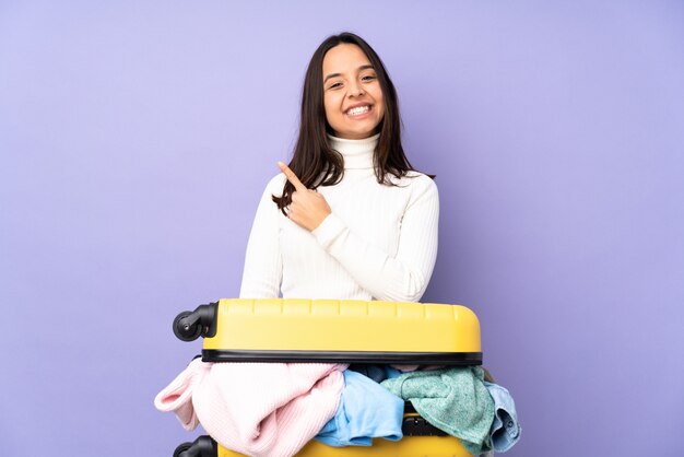 Viajero joven con una maleta llena de ropa apuntando hacia un lado para presentar un producto