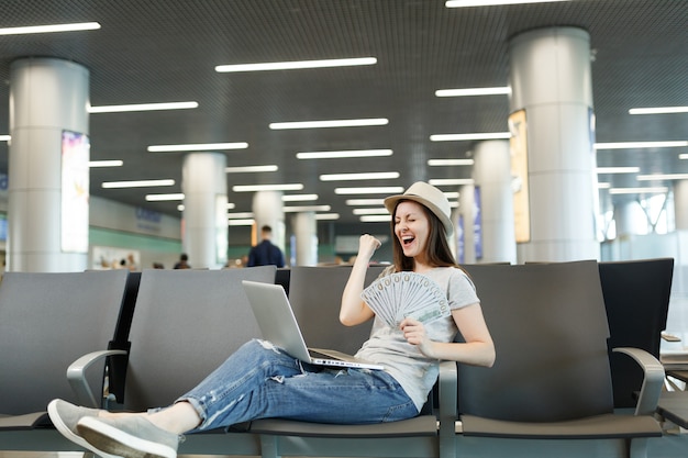 Viajero lleno de alegría mujer turista que trabaja en la computadora portátil mantenga un paquete de dólares en efectivo, haga el gesto del ganador, espere en el vestíbulo del aeropuerto