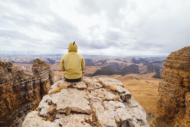 Un viajero masculino con una chaqueta se sienta al borde de un acantilado y disfruta de la naturaleza de la montaña