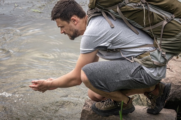 Un viajero masculino con una gran mochila de senderismo bebe agua de un río de montaña.