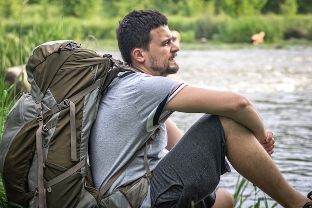 Un viajero masculino con una gran mochila de senderismo descansa cerca del río.